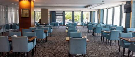 A spacious dining area with blue chairs and tables, patterned carpet, and large windows offering views of the outdoors.