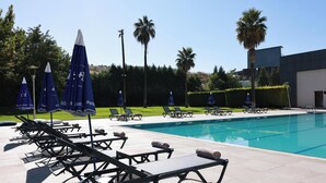 Uma área na beira da piscina com espreguiçadeiras e guarda-sóis, piscina azul cristalina e palmeiras.