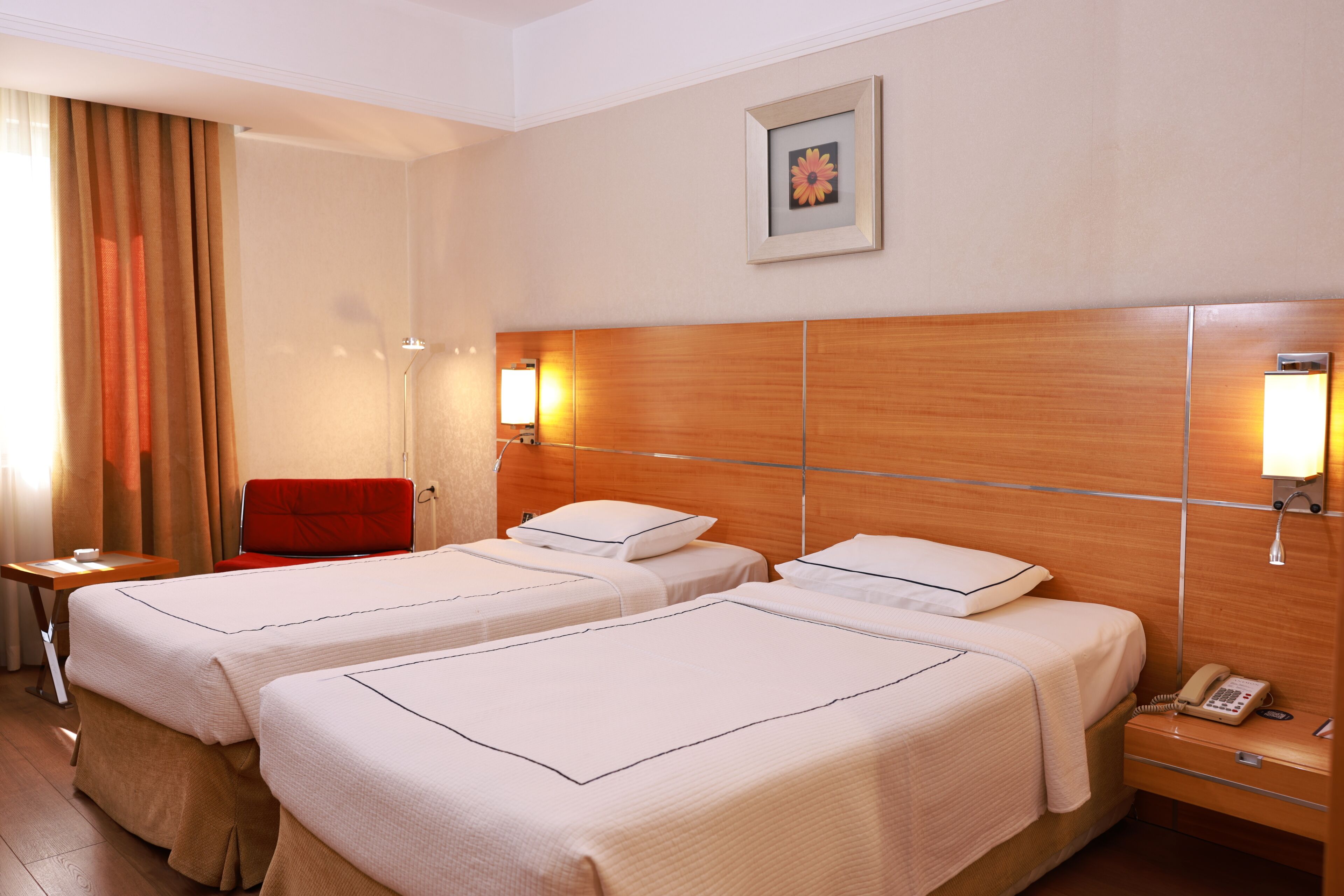 A hotel room with two single beds, a wooden headboard, bedside lamps, a red chair, a small table, and a telephone on the nightstand.