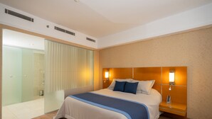 Quarto de hotel moderno com uma cama grande, uma mesa de cabeceira, divisória de chuveiro em vidro e piso de madeira.