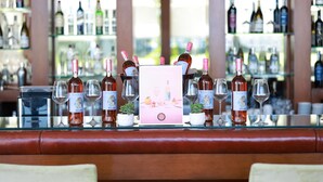 Uma área de bar com garrafas e copos dispostos sobre uma mesa de vidro, com uma janela ao fundo.
