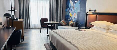 Habitación de hotel con cama, escritorio, silla, una mesita y un ventanal con cortinas.