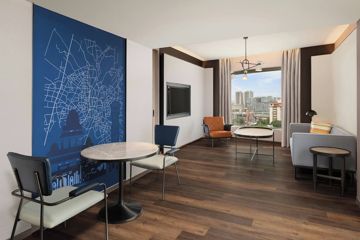 Une chambre d’hôtel moderne avec vue sur la ville, une table à manger ronde, un canapé et un téléviseur fixé au mur.