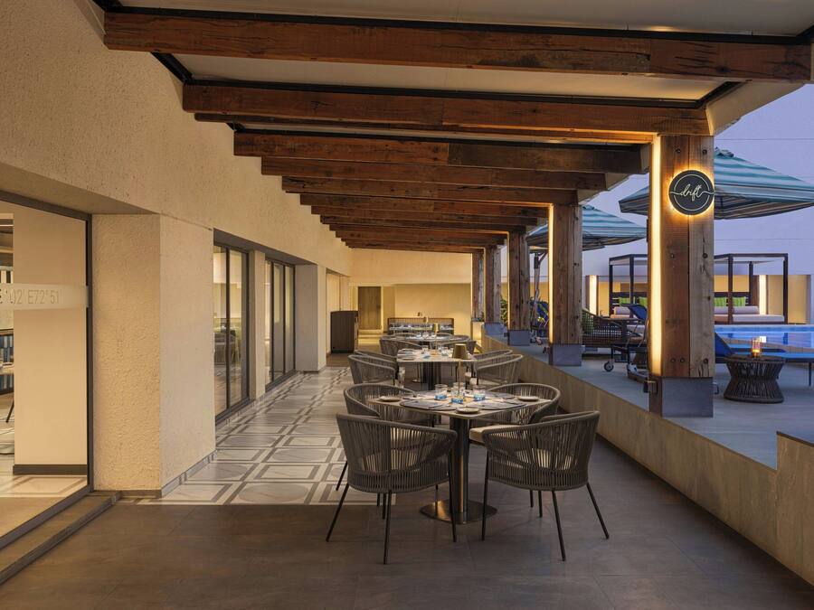 An outdoor dining area with wooden beams, tables, and chairs, and a pool in the background.