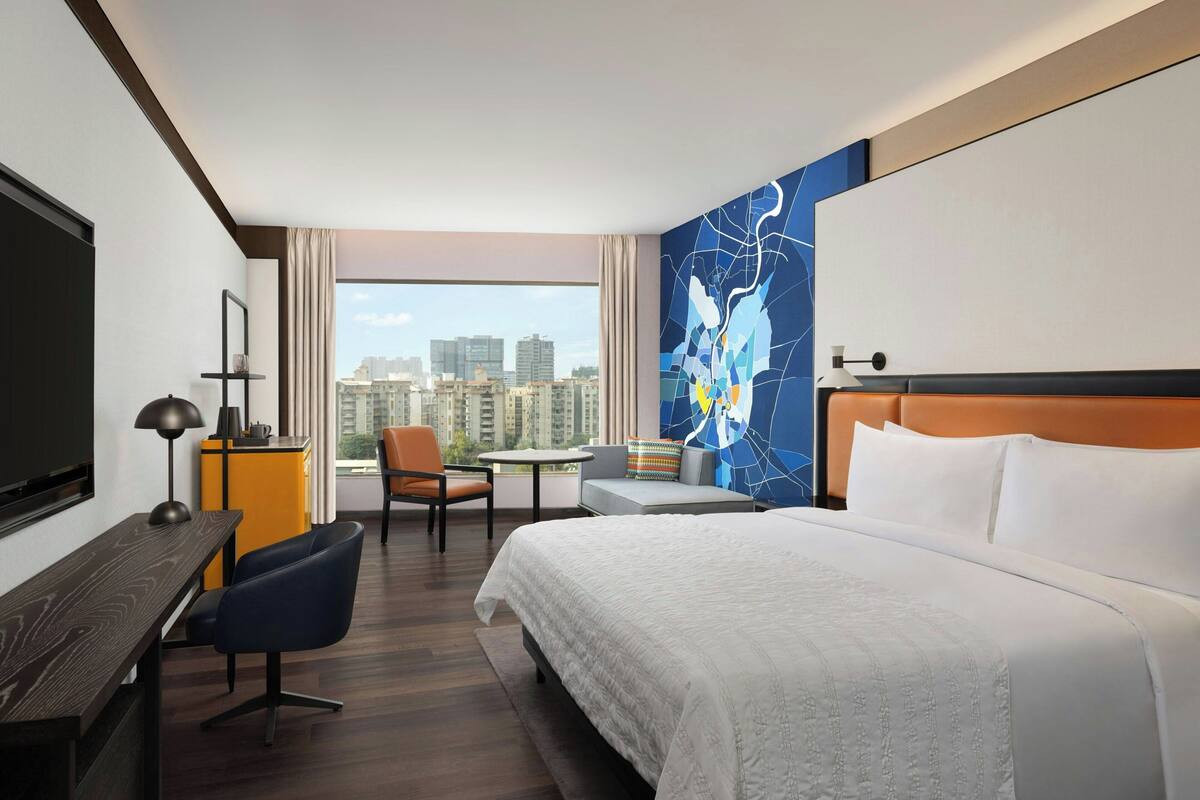 Une chambre d’hôtel avec un grand lit, un bureau, une chaise, une petite table et une vue sur les toits de la ville.