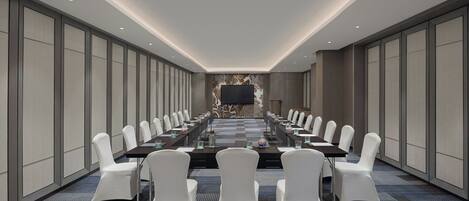 A conference room with a long table, white chairs, and a large screen at the front.