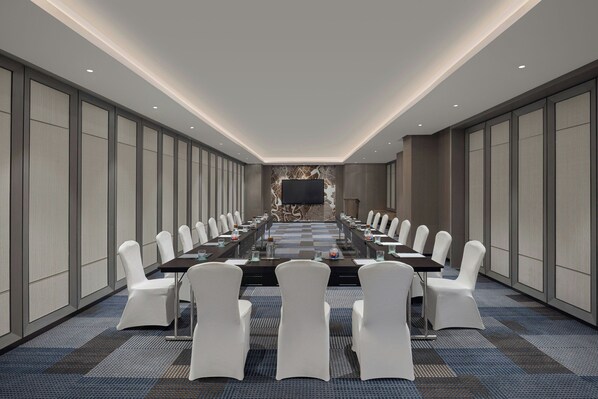 Un salón de conferencias con una larga mesa, sillas blancas y una pantalla grande al frente.