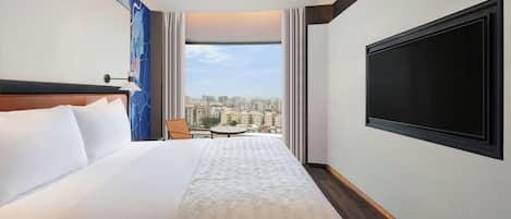 Quarto de hotel com uma cama grande, uma TV de tela plana na parede, vista da cidade através de uma janela ampla e uma pequena mesa com uma cadeira.