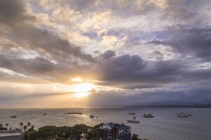 Un paisaje urbano costero con un atardecer sobre el mar, que muestra varias embarcaciones y un edificio destacado con techo azul.