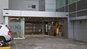 Entrada de un edificio moderno con puerta de vidrio, un coche blanco estacionado en frente y una persona entrando.