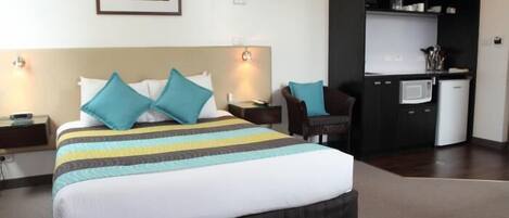 A hotel room with a bed, a nightstand, a desk with a chair, a microwave, and a built-in kitchen cabinet.