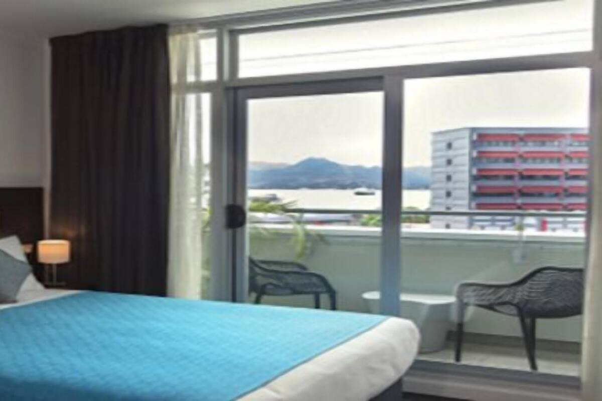 Habitación de hotel con cama, dos cuadros en la pared, ventana con vistas a edificios y montañas, y balcón con sillas.