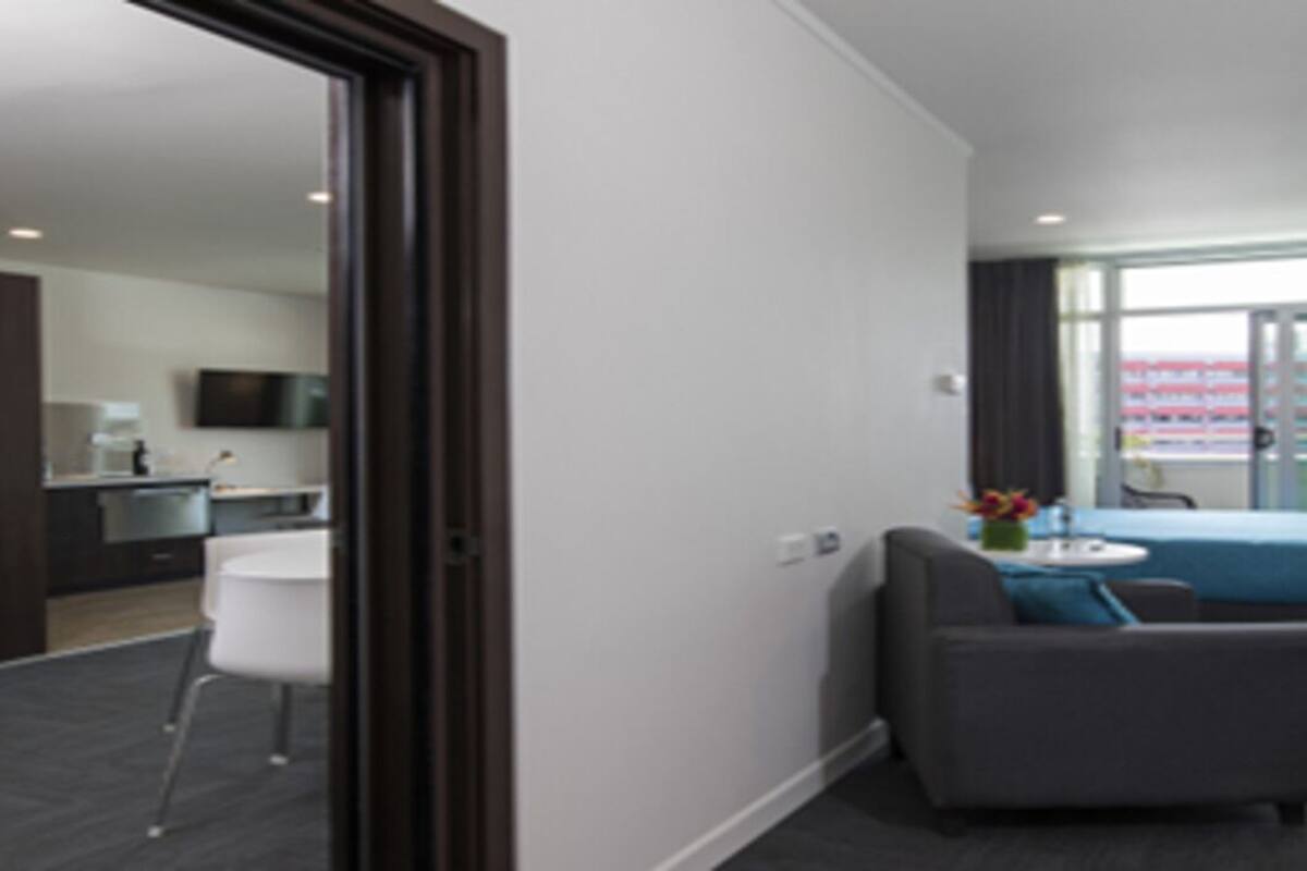 Habitación de hotel con cama, escritorio, silla y una pequeña mesa con flores. Se aprecia una ventana con cortinas y vistas al exterior.