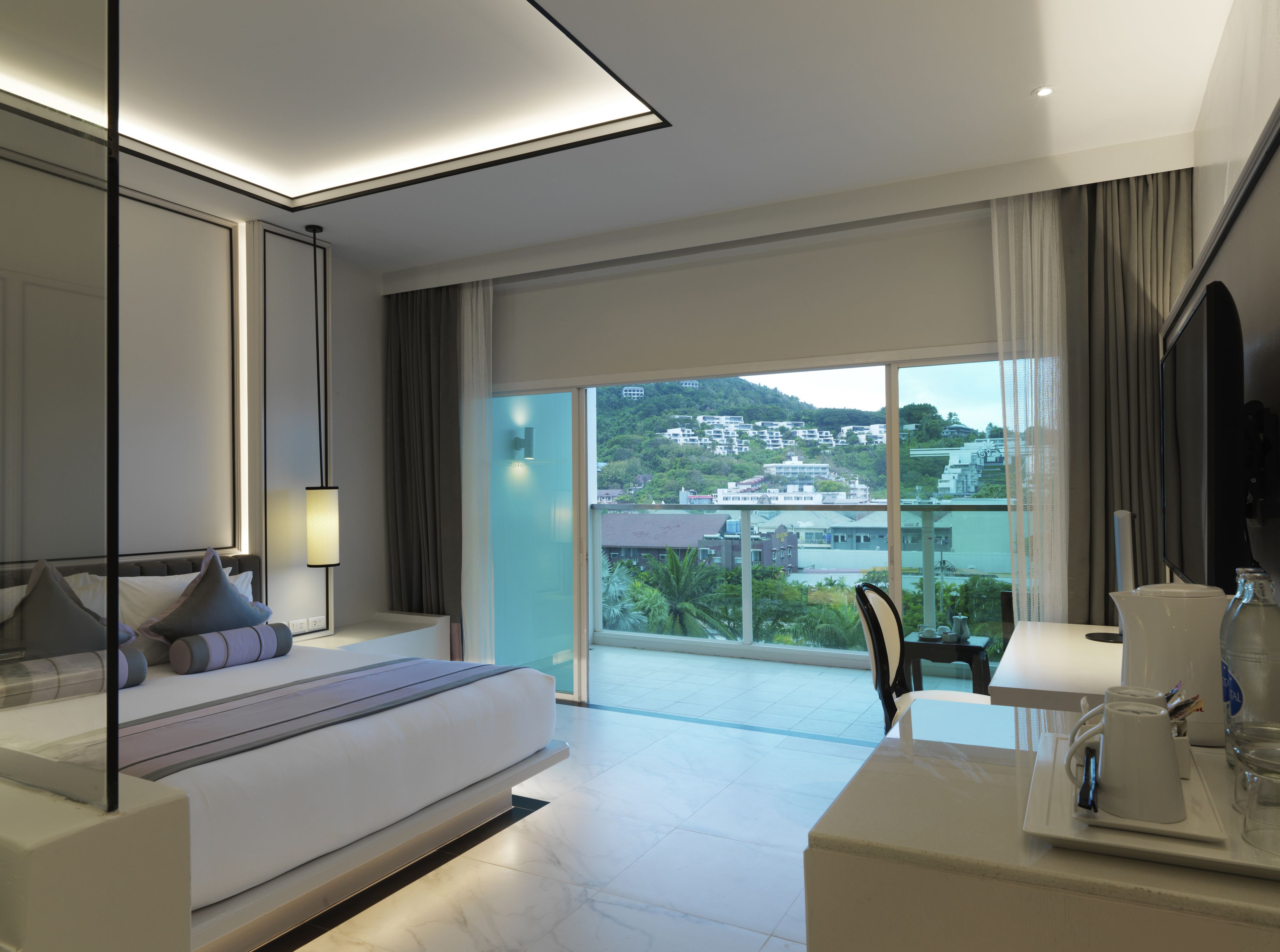 Una habitación de hotel moderna con una cama grande, un área de descanso, un balcón con vistas a la ciudad y un baño con tocador y espejo.
