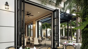 Un área de comedor al aire libre con mesas redondas y sillas de mimbre, conectada a un espacio interior por puertas de cristal.