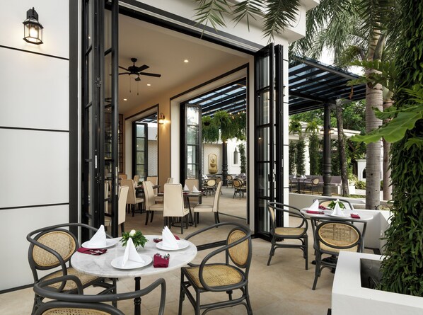 An outdoor dining area with round tables and wicker chairs, connected to an indoor space by glass doors.