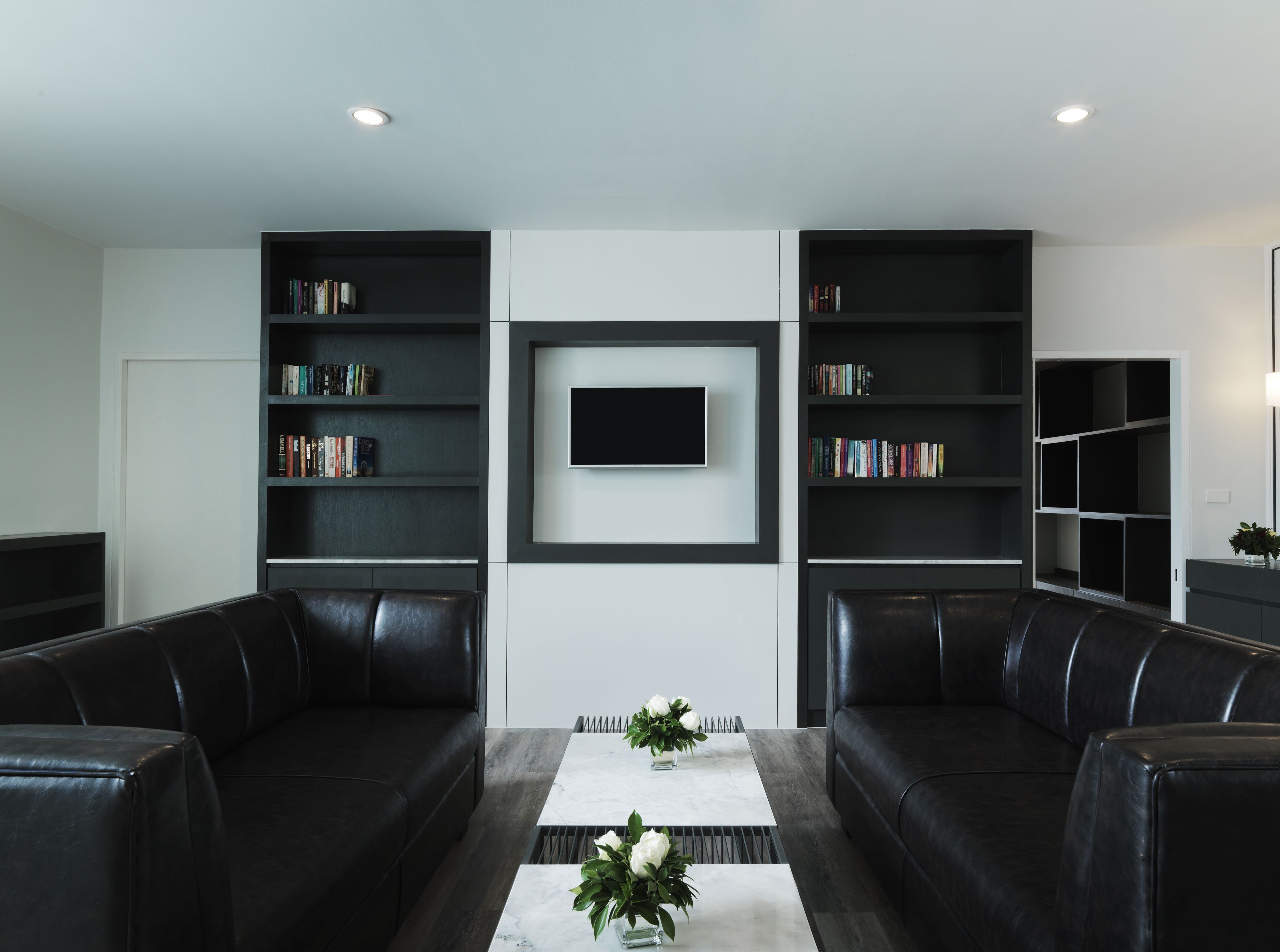 A modern living room with two black leather sofas, a central coffee table with a vase of flowers, and built-in bookshelves on either side.