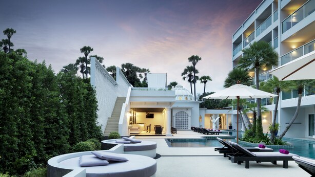Zona de piscina moderna con sillones de descanso, una piscina y un edificio de fachada blanca con palmeras.