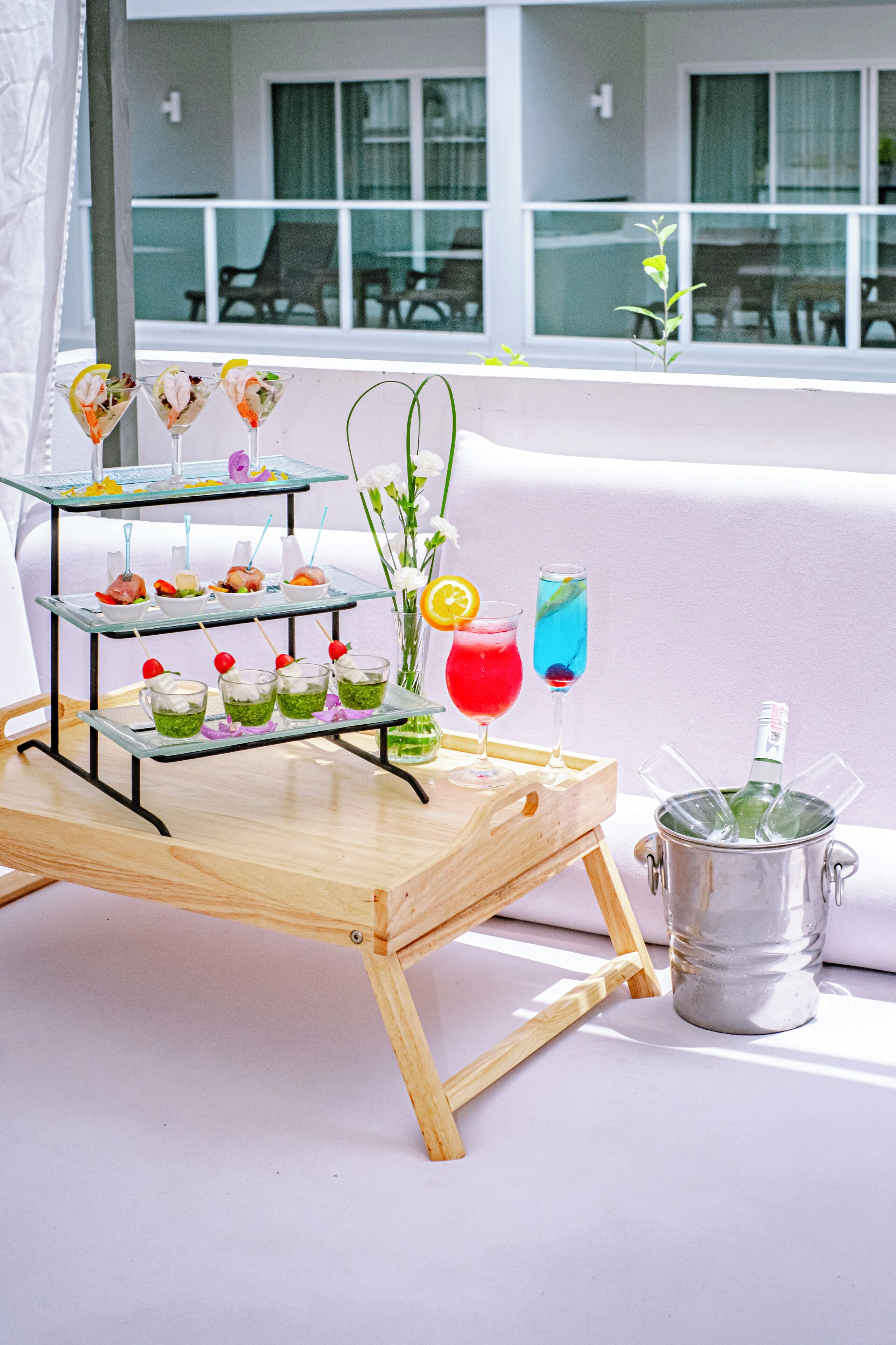 A glass display stand with various drinks and appetizers on a wooden table, a vase with flowers, and a bottle of champagne in a metal bucket.