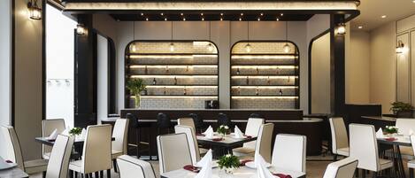 A modern restaurant with a long bar, white chairs, and a brick wall display.