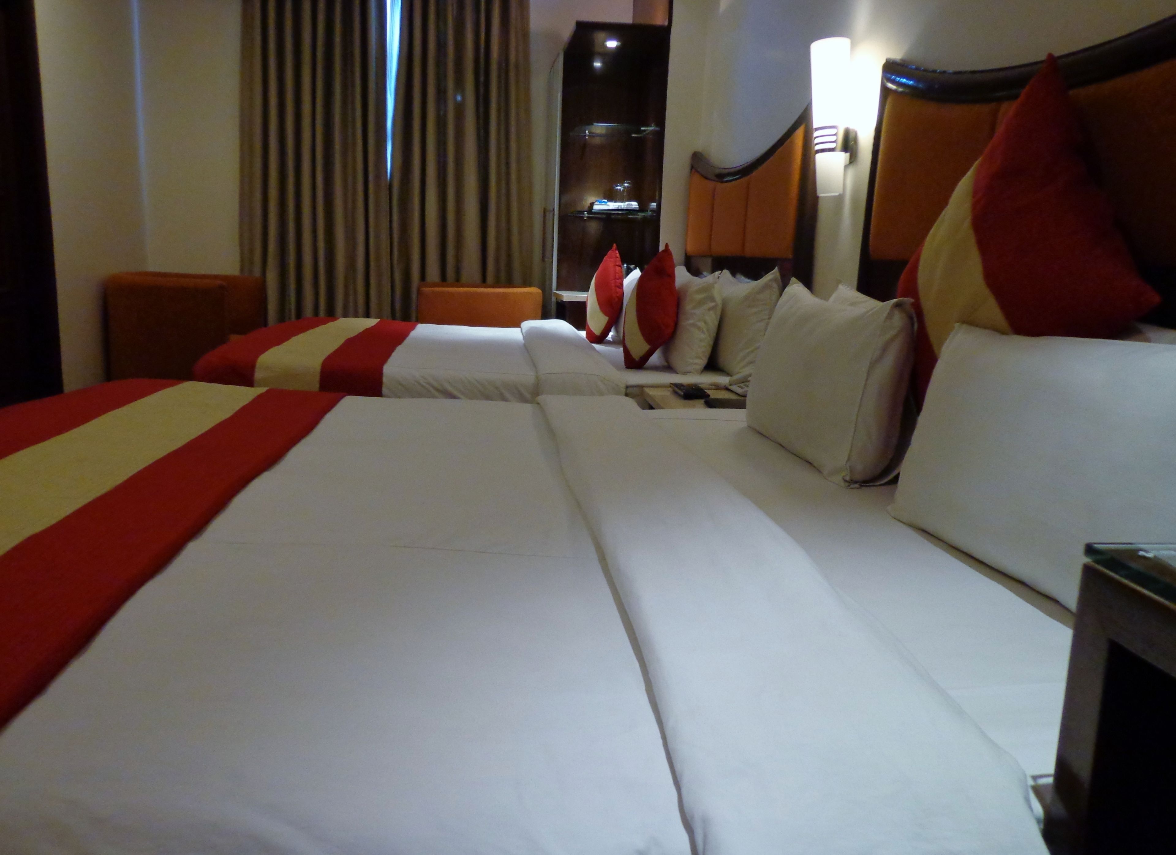 A hotel room with three beds, each with white linens and red accents. There is a wooden headboard, a nightstand with a lamp, and a window with curtains.
