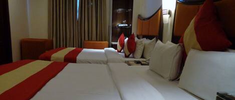 Habitación de hotel con tres camas, cada una con ropa blanca y detalles rojos. Cuenta con un cabecero de madera, una mesita de noche con lámpara y una ventana con cortinas.