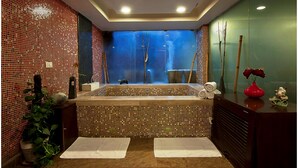 Un baño con una bañera grande, un espejo y un lavabo. Hay toallas en el suelo y una planta al lado del lavabo.