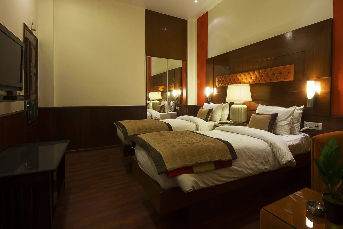 A hotel room with two beds, a desk, a chair, a plant, and a mirror.