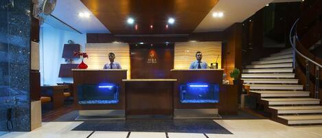 A reception desk with two receptionists, a wooden reception area, a fish tank, and a staircase.