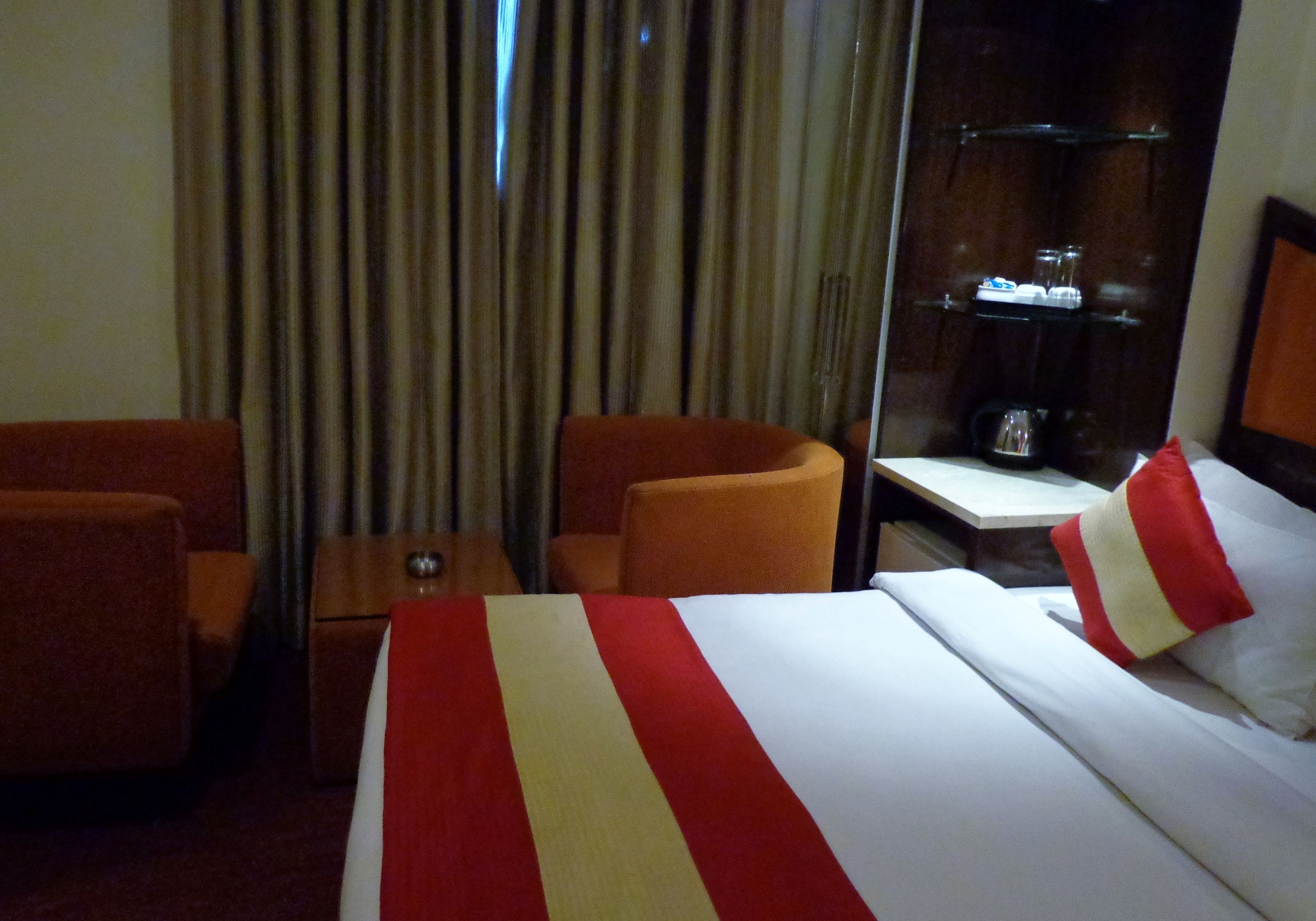 Quarto de hotel com uma cama, duas cadeiras, uma mesinha, um espelho e uma prateleira com produtos de higiene pessoal.