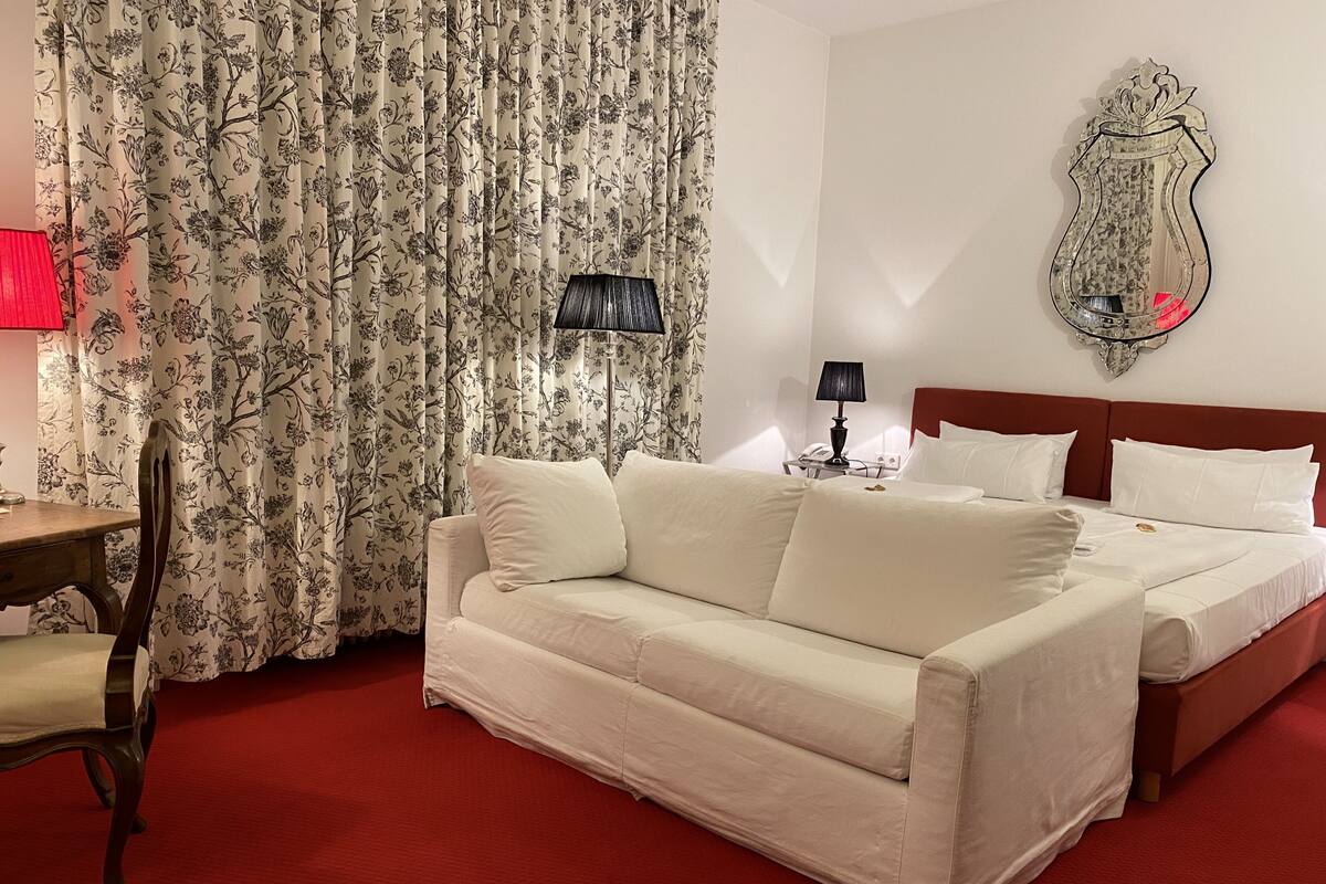 Habitación de hotel con cama, sofá, alfombra roja, lámpara y cortinas estampadas con motivos florales.