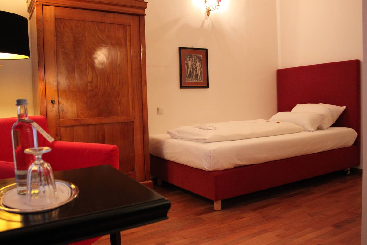 Una habitación con un armario de madera, una cama con ropa blanca, una silla roja y una mesa con una botella y vasos.