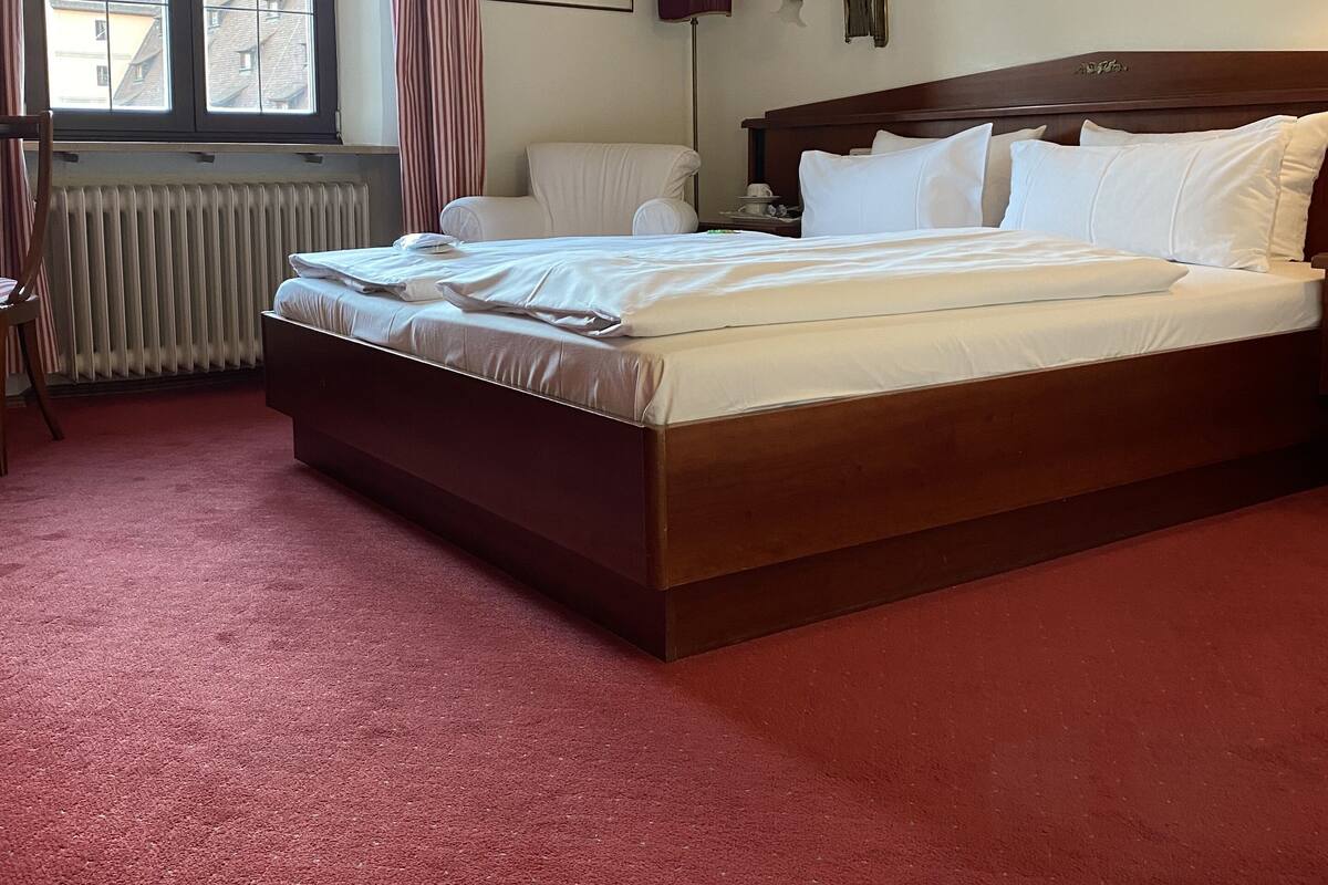 Una habitación de hotel con una cama grande, un radiador, una ventana con cortinas y cuadros en la pared.