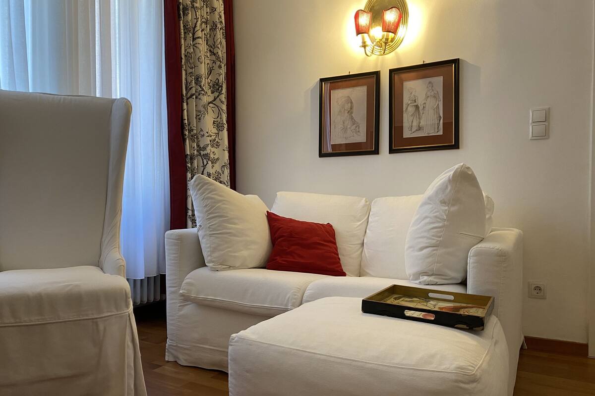 Una sala de estar con un sofá blanco, un cojín rojo y una mesa de centro con un libro. Hay dos obras de arte enmarcadas en la pared y una ventana con cortinas.