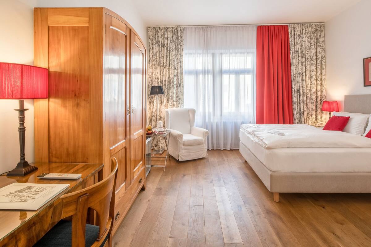 Habitación de hotel con un armario de madera, un escritorio con lámpara, un sillón blanco, una cama con almohadas rojas y un ventanal con cortinas de tul y un lazo rojo.