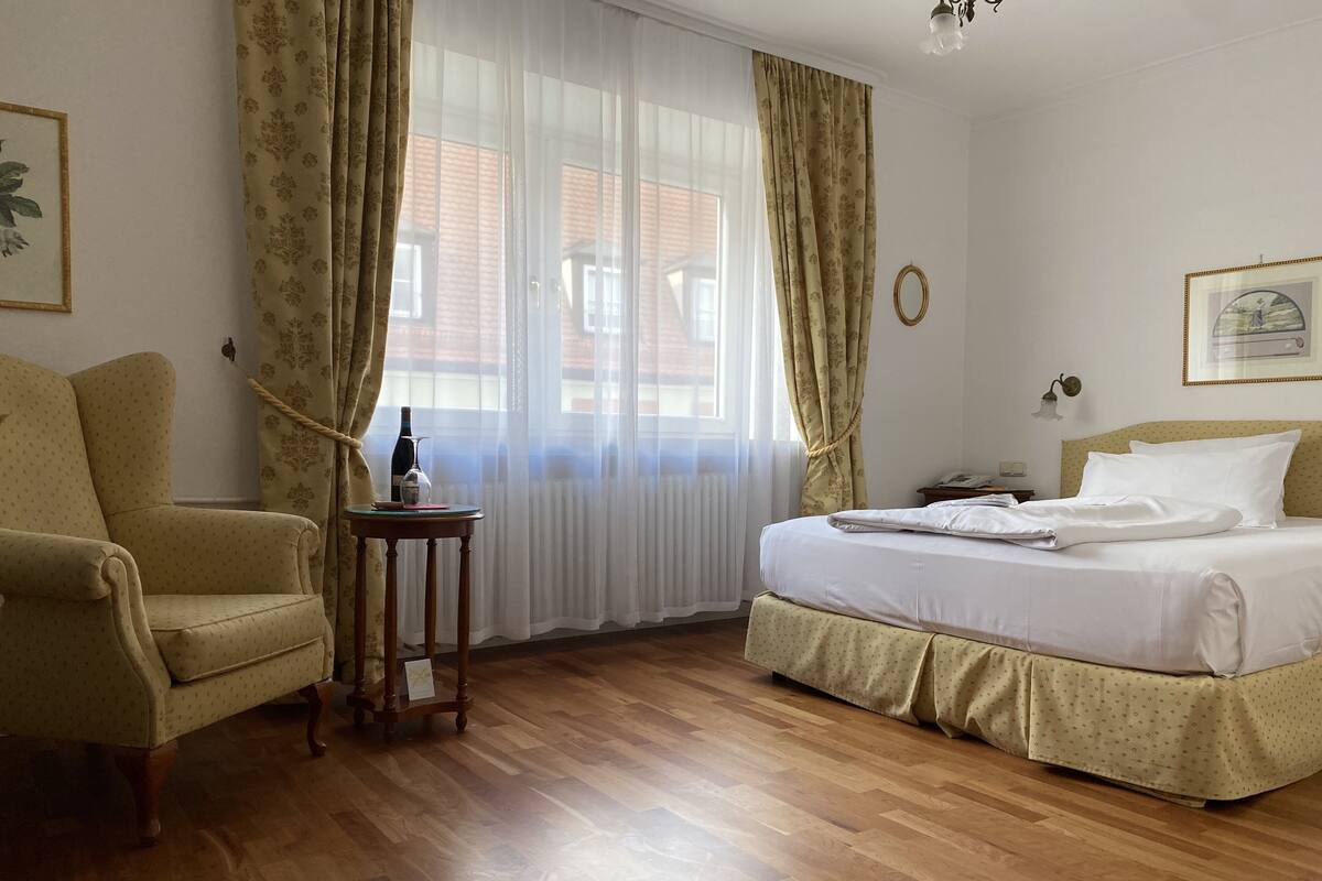 Una habitación de hotel con cama, una silla, una mesita con una botella y vasos, y una ventana con cortinas.