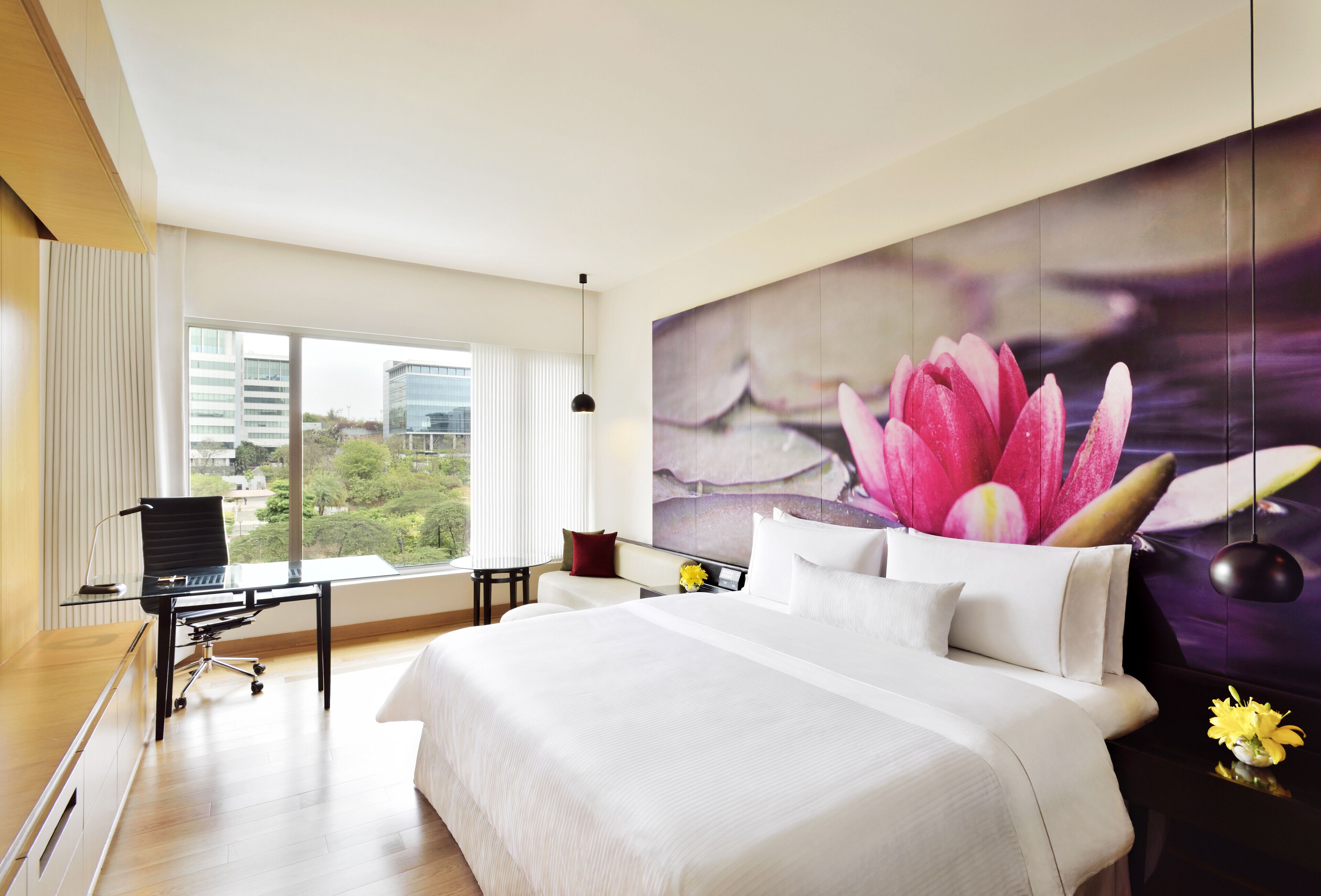 A modern hotel room with a large bed, a desk with a chair, and a view of greenery through large windows.
