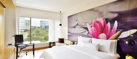 A modern hotel room with a large bed, a desk with a chair, and a view of greenery through large windows.