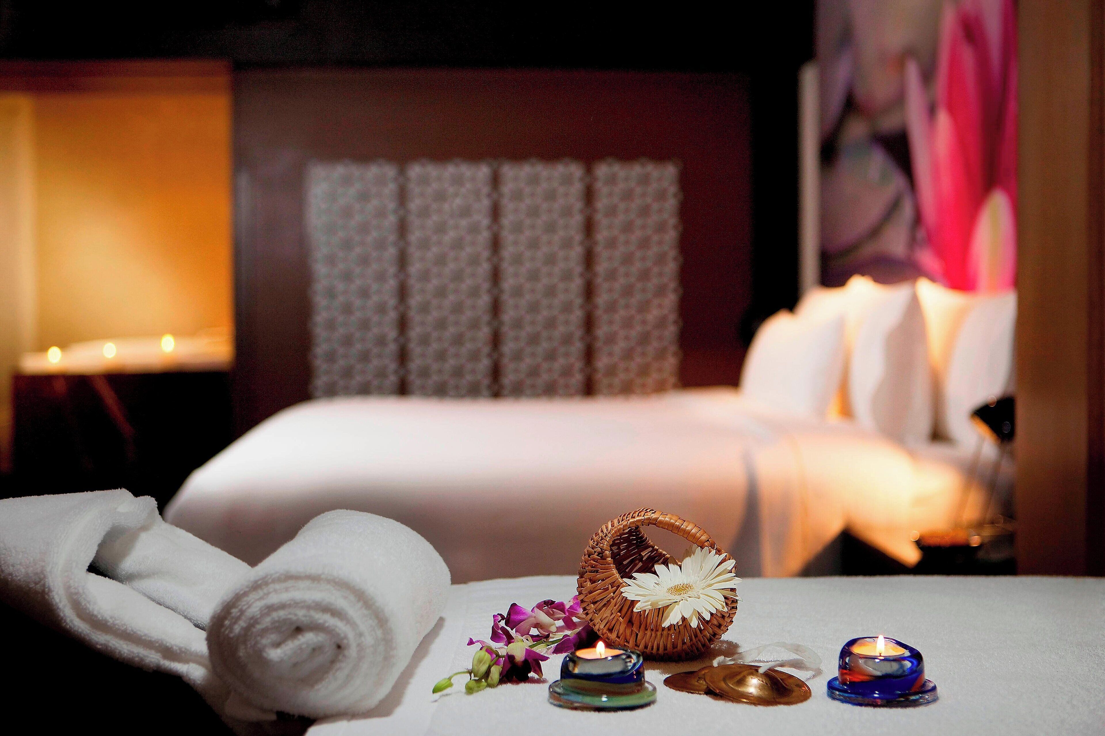 A neatly made bed with a white comforter, a rolled white towel, a small wicker basket with a white flower, and two candles on a bedside table.