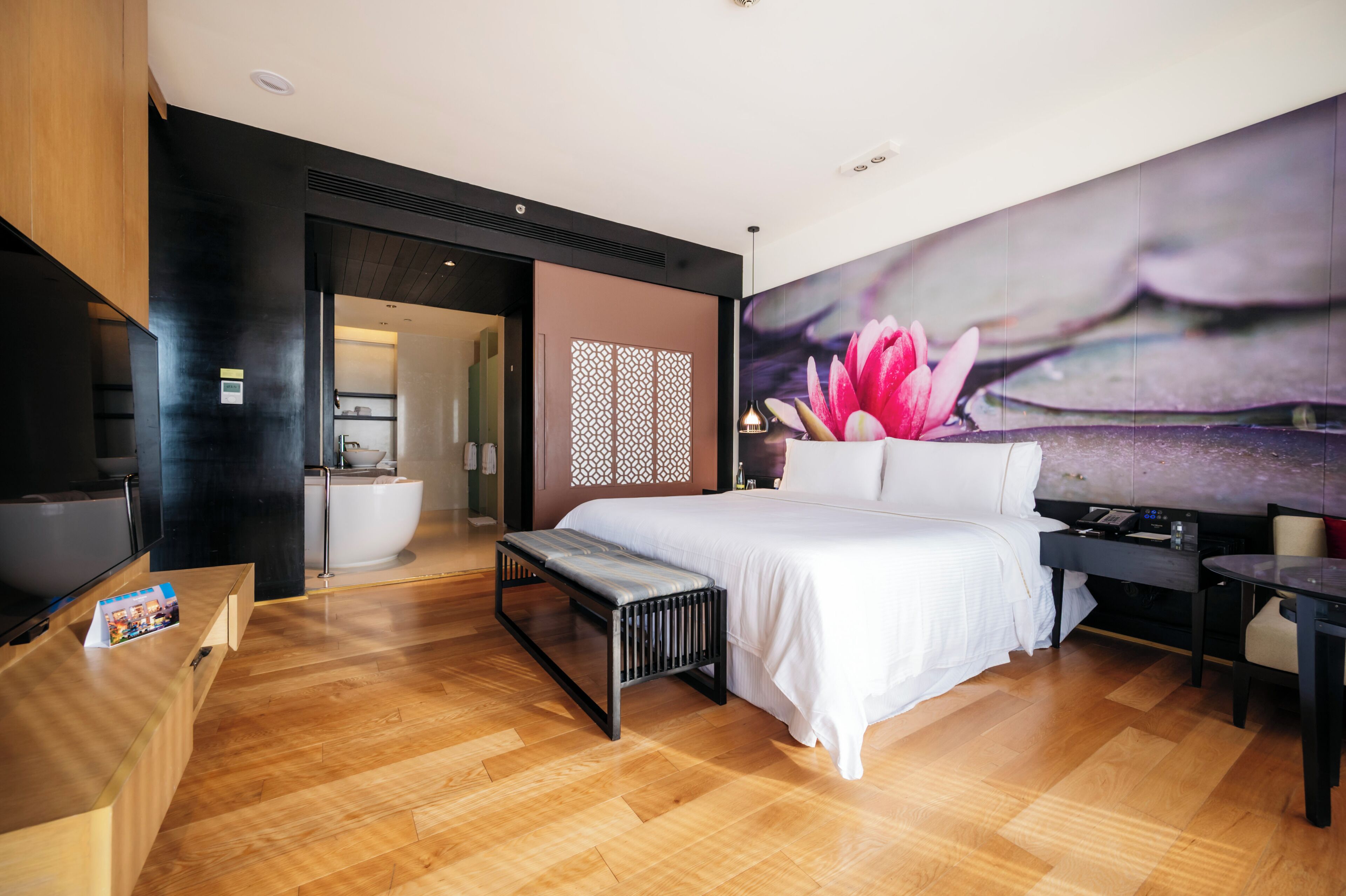 Una camera d'albergo moderna con un letto grande, una panca di legno, una scrivania e un bagno parzialmente visibile attraverso una porta aperta.