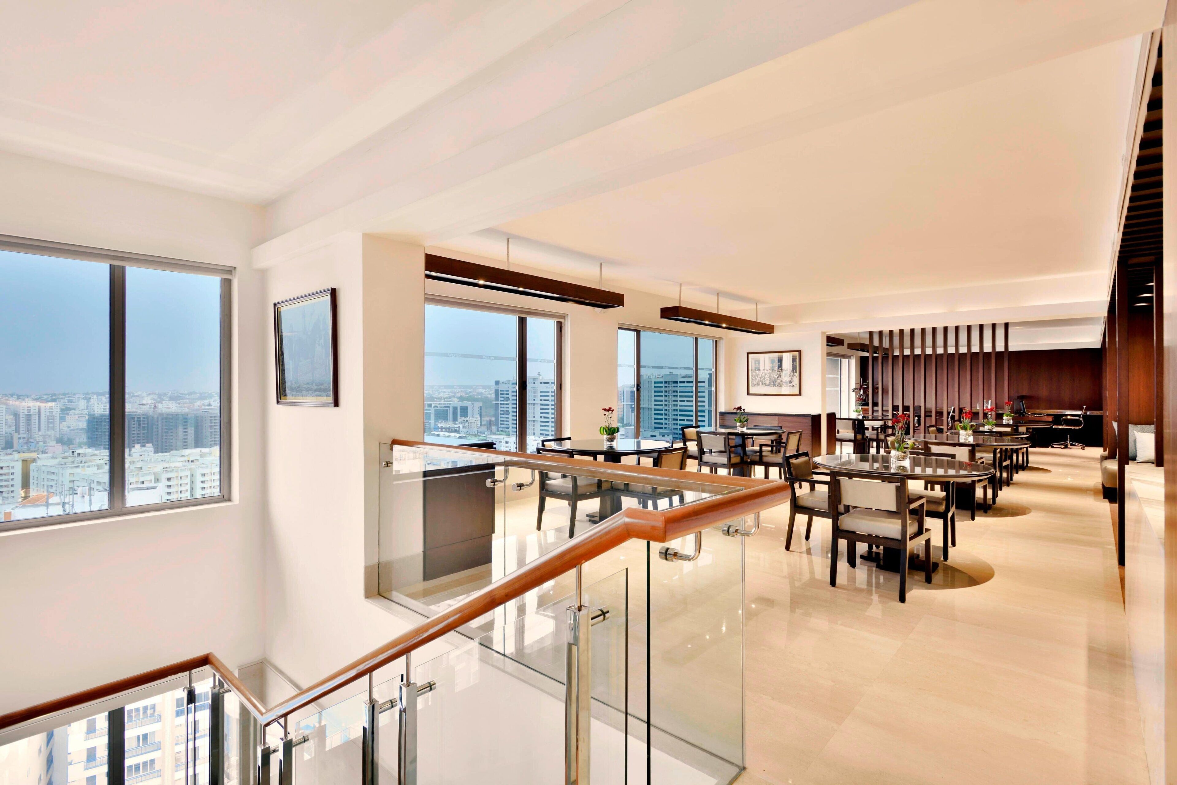 A spacious hotel lobby with a high ceiling, large windows offering city views, a central seating area, and a staircase with glass railings.