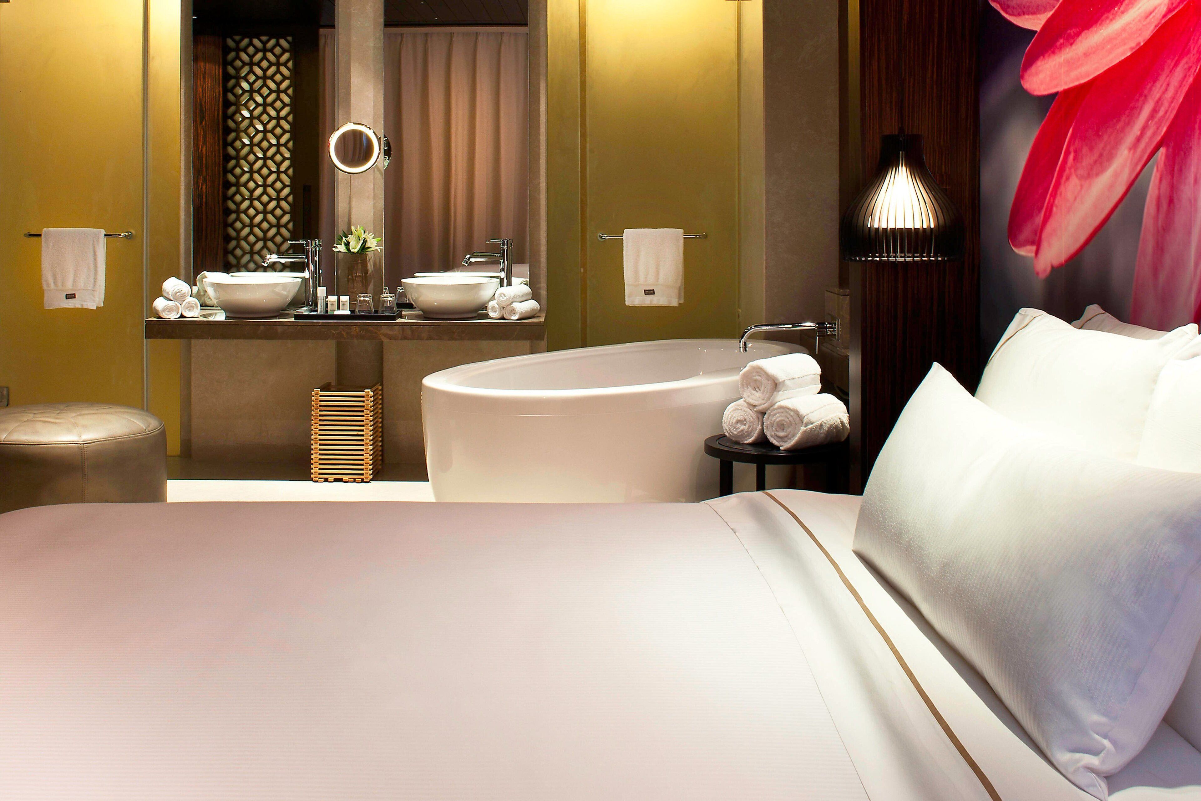Una habitación de hotel moderna con una cama grande, un espejo redondo, una bañera y un tocador con lavabo y toallero.