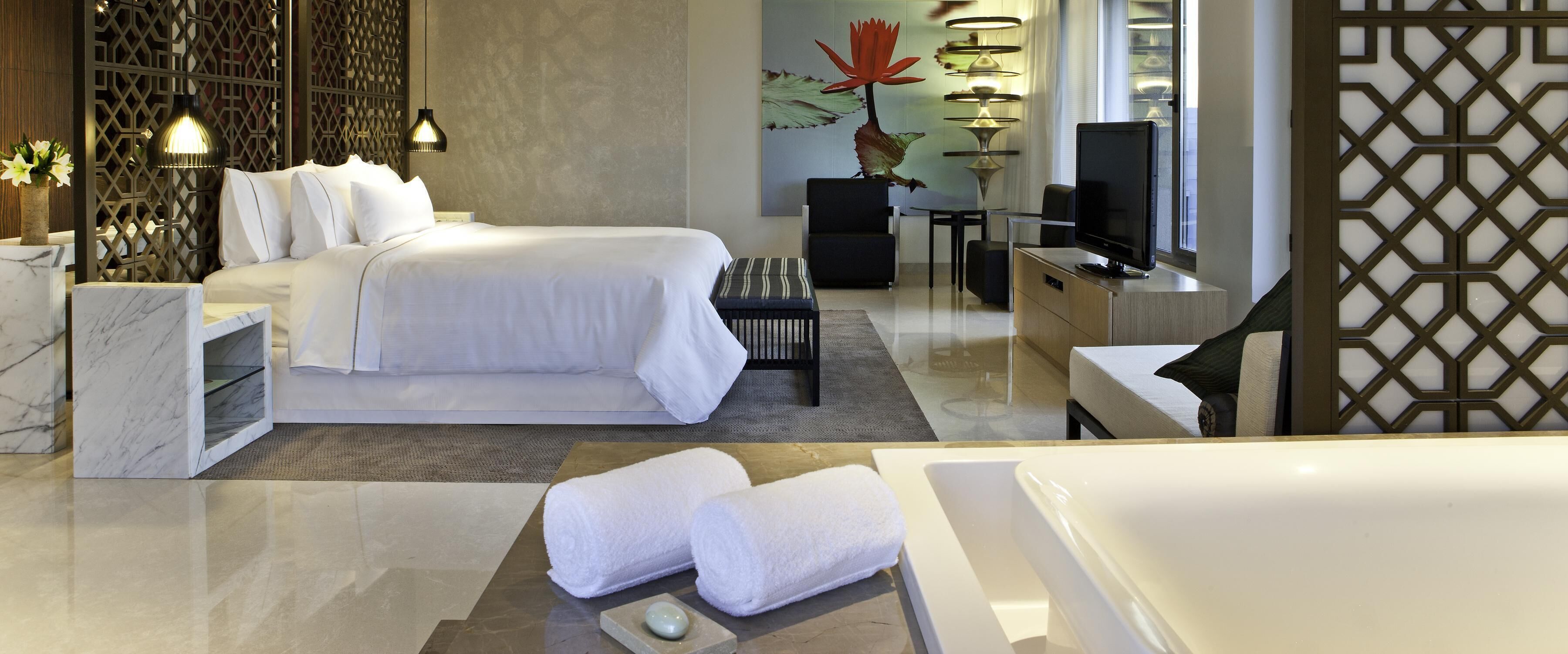 A modern hotel room with a large bed, a desk, a chair, a TV, and a bathtub.