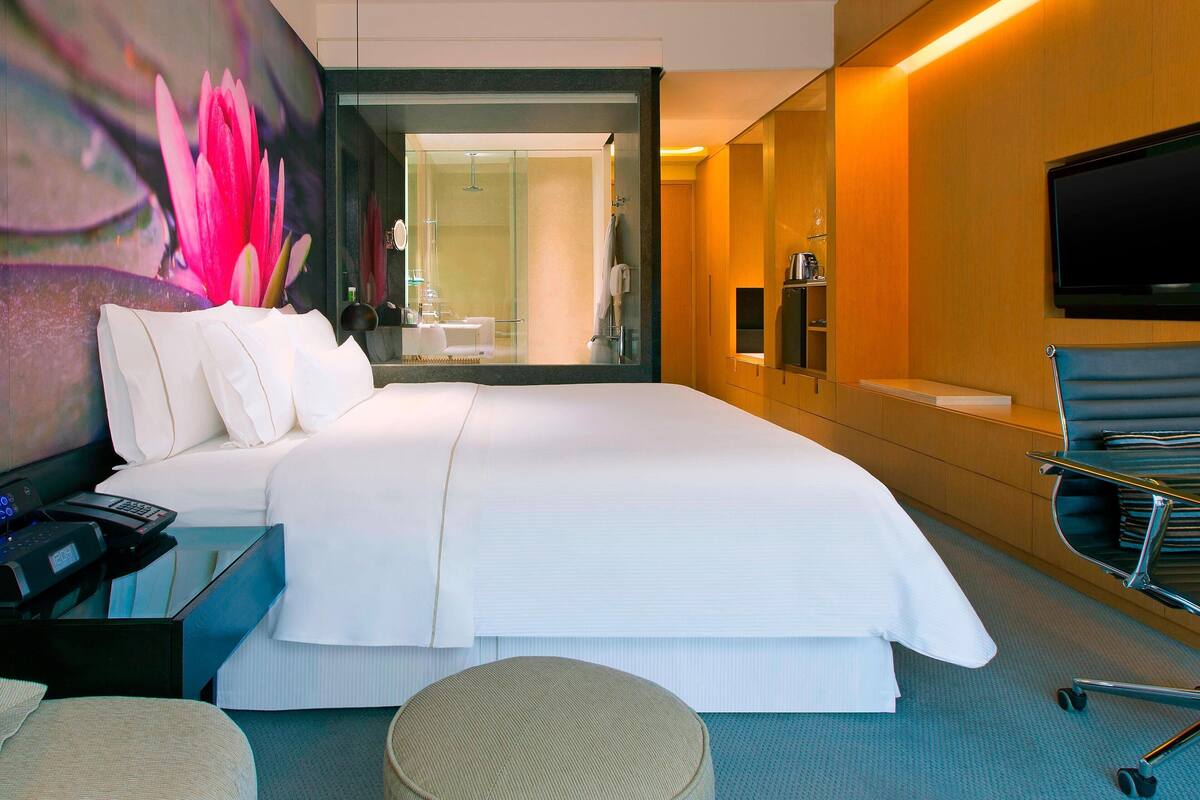 A modern hotel room with a large bed, a desk with a computer, a flat-screen TV, and a bathroom visible through a glass door.