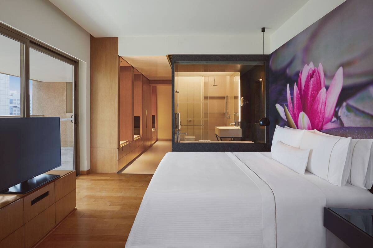 A modern hotel room with a large bed, a flat-screen TV, wooden flooring, and a view of the city through a large window.