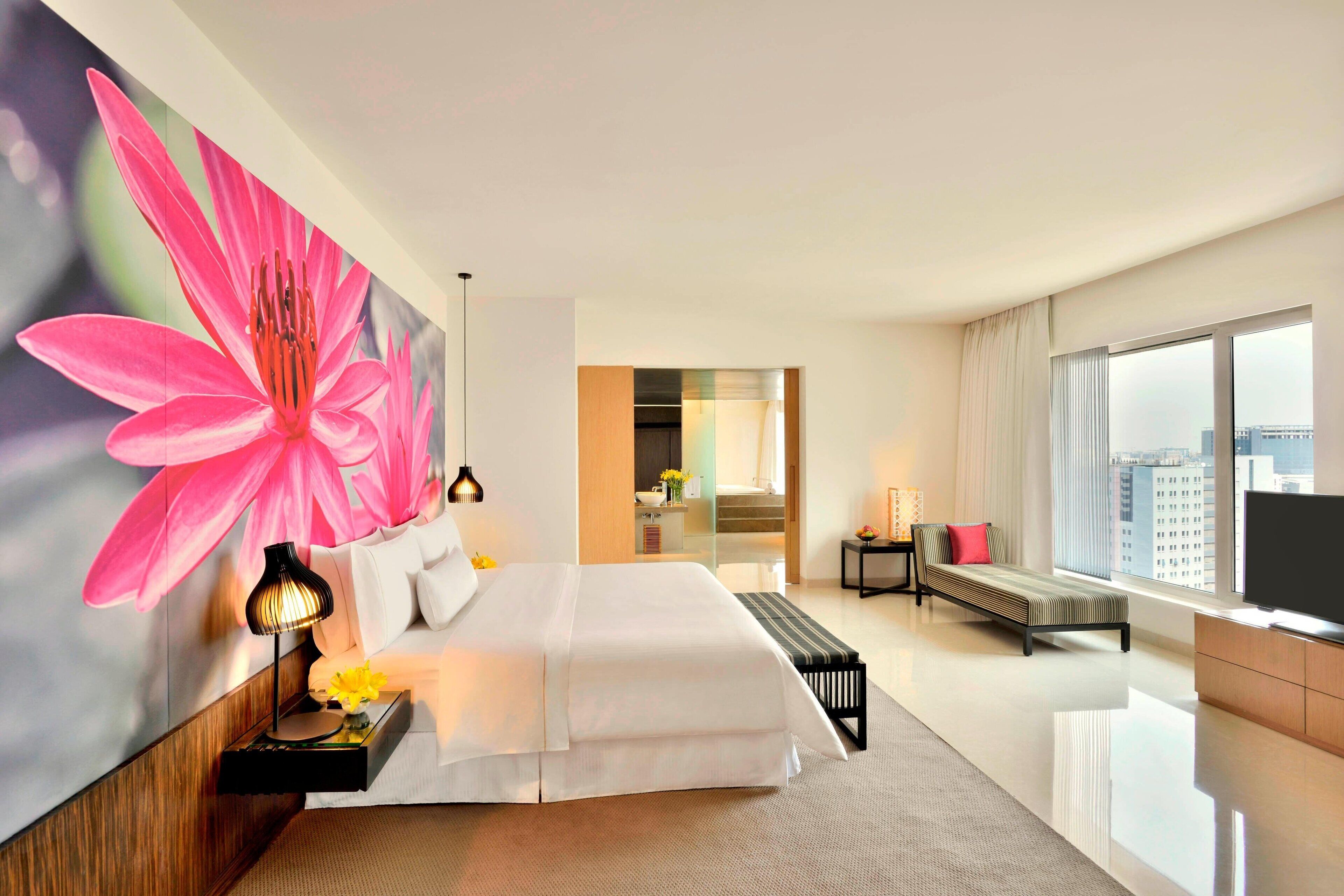 A modern hotel room with a large bed, a TV, a bedside table with a lamp, and a large window with a city view.