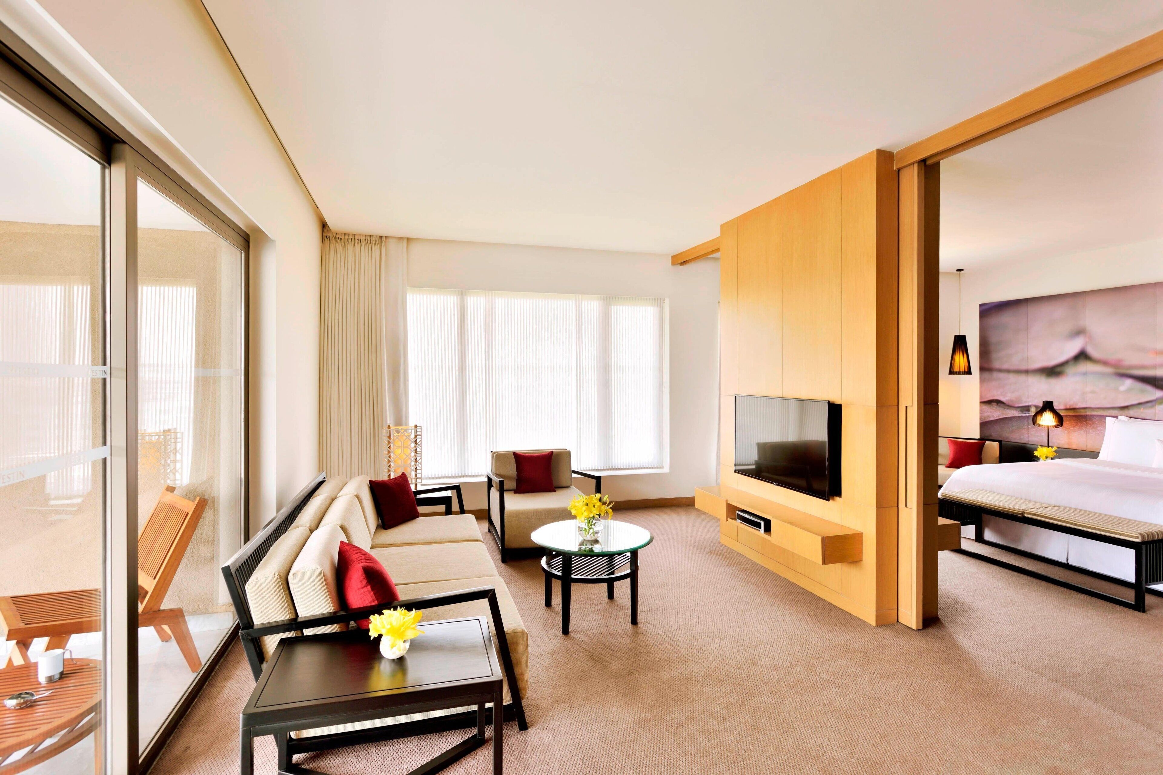 A hotel room with a sofa, a small table with flowers, a TV, a bed, and a balcony with a chair.