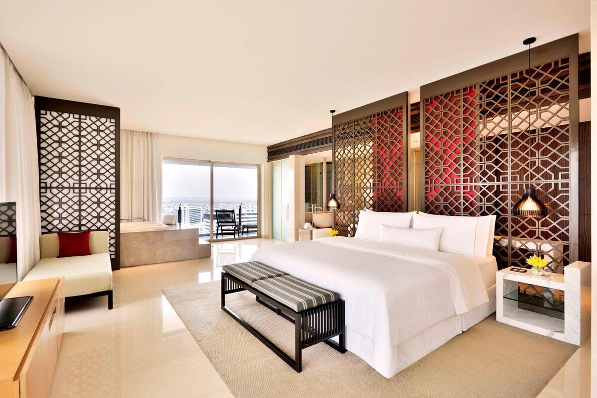 A modern hotel room with a large bed, a desk, a bench, and a view of the city.