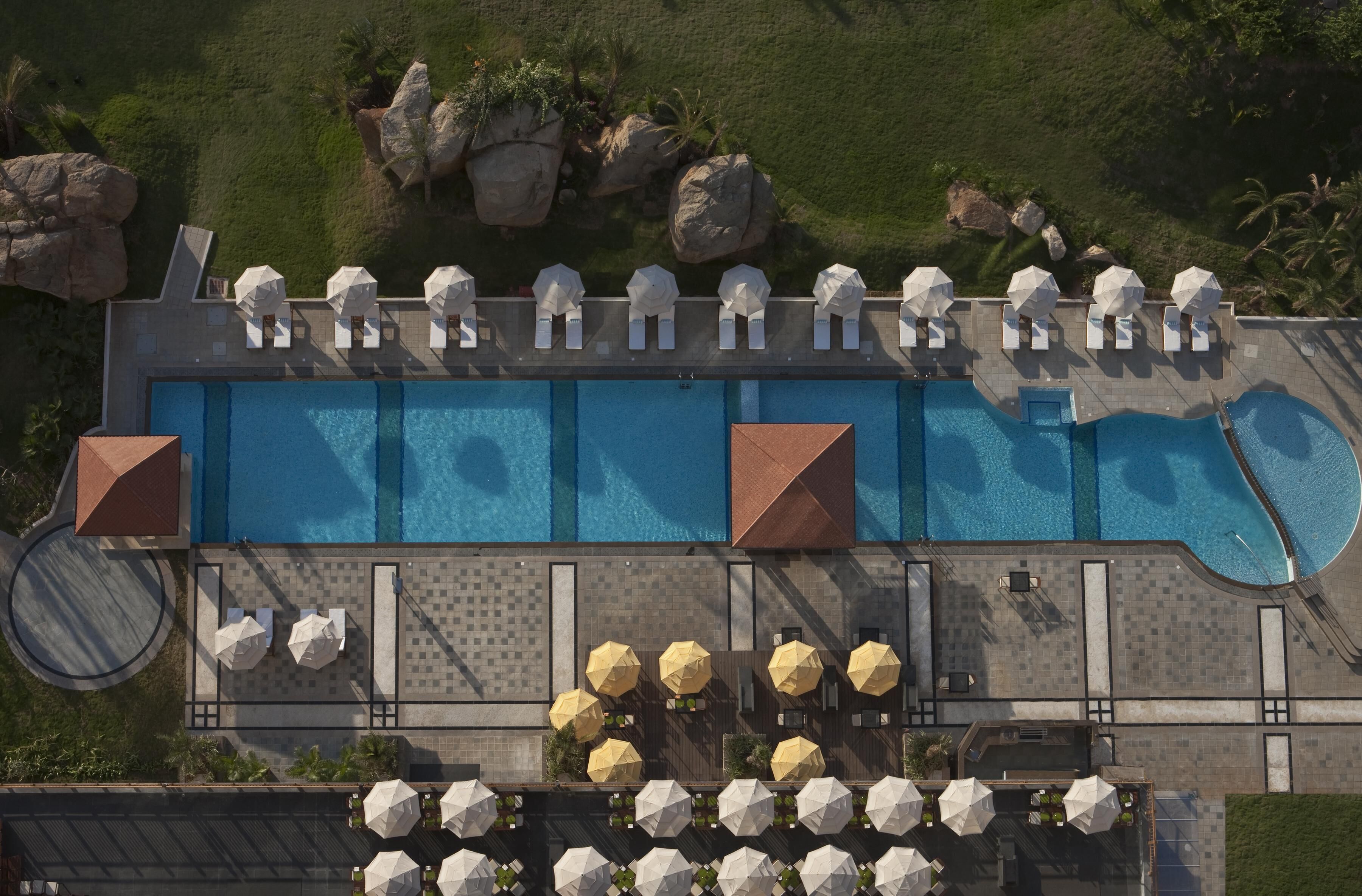 Vue aérienne d’un espace piscine d’hôtel, avec de nombreux transats et parasols, une grande piscine et une végétation luxuriante tout autour.
