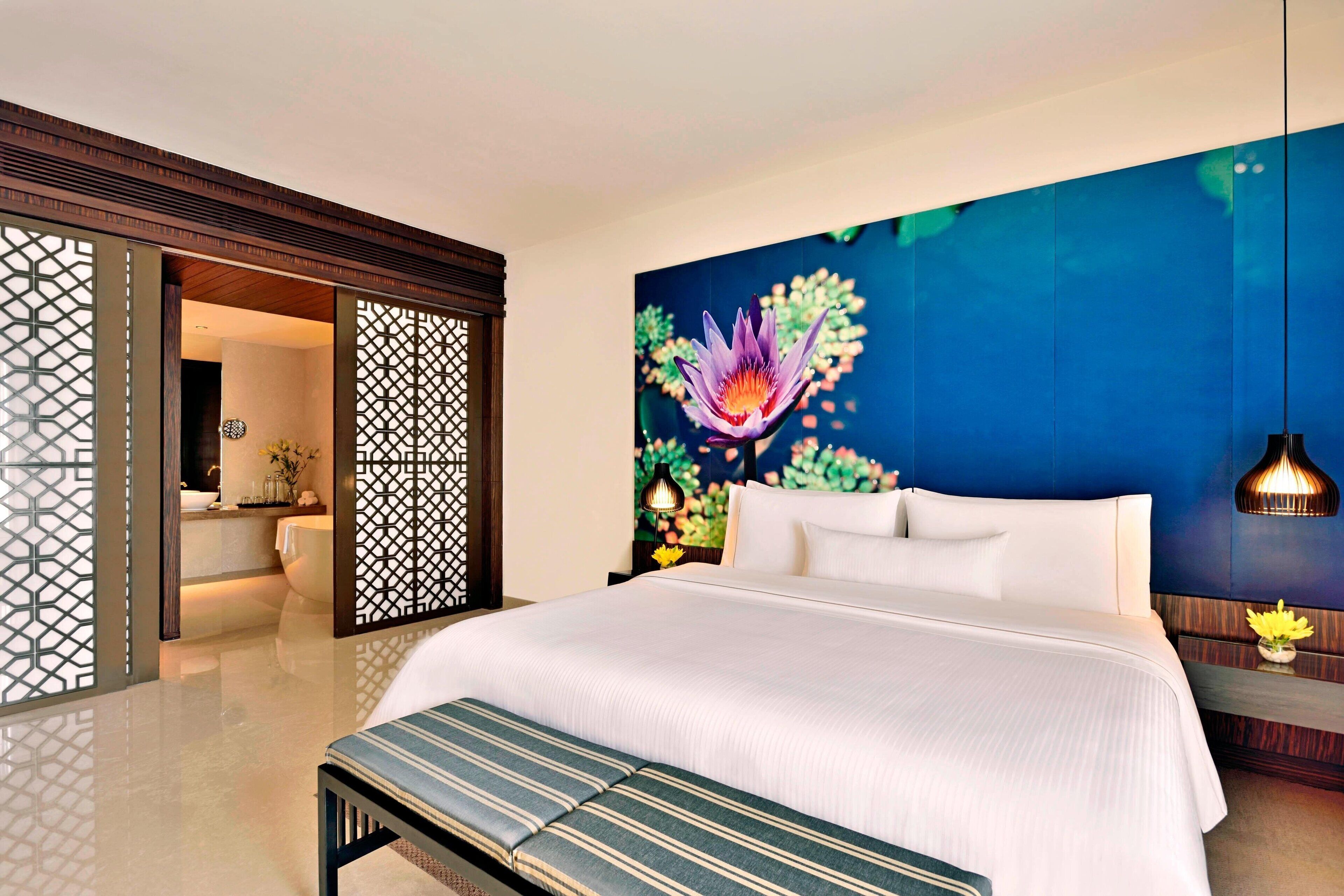 A hotel room with a large bed, a bench, a wall-mounted lamp, and a decorative wall with a floral painting.
